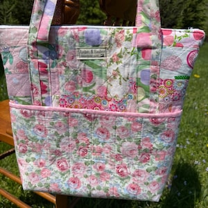 May include: A quilted tote bag with a floral pattern in shades of pink, white, and green. The bag features a zippered top, a pink pocket, and matching handles. The label reads "Arden & Ember".