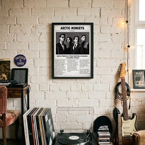 A framed black-and-white poster featuring the band Arctic Monkeys in a newspaper layout hangs on a white brick wall above a record player and an electric guitar.