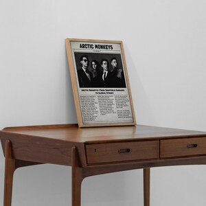 A framed vintage-style newspaper poster featuring the band Arctic Monkeys leans against a plain white wall on top of a mid-century modern wooden desk.