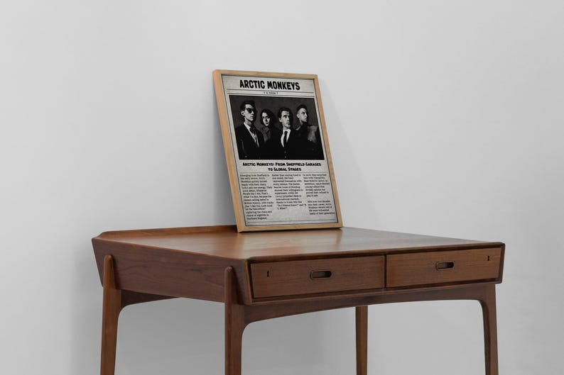 A framed vintage-style newspaper poster featuring the band Arctic Monkeys leans against a plain white wall on top of a mid-century modern wooden desk.