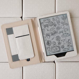 Op de afbeelding: Een Kindle e-reader in een lichtbeige hoes, open om een scherm te tonen met zwart-witte illustraties met strandthema. De hoes ligt op een achtergrond van bijpassende beige vierkanten.