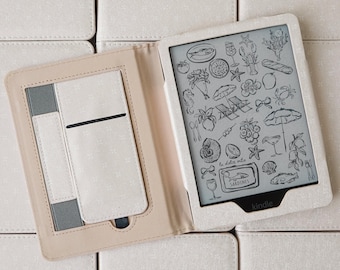 Könnte beinhalten: Ein Kindle E-Reader in einer hellbeigen Hülle, geöffnet, um einen Bildschirm mit schwarz-weißen Illustrationen zum Thema Strand zu zeigen. Die Hülle liegt auf einem Hintergrund aus ähnlichen beigen Quadraten.