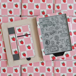 Op de afbeelding: Een Kindle e-reader hoes met een roze en wit geblokt patroon en rode aardbei-illustraties. De binnenzijde heeft een beige voering, een grijze elastische band en een kaartsleuf. Het scherm toont zwart-witte lijntekeningen van diverse iconen.