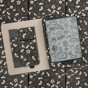 Könnte beinhalten: Eine dunkelgraue Kindle-E-Reader-Hülle mit weißem Blumenmuster. Das Innere ist hellbeige mit einem passenden Kartenfach und einem elastischen Band. Der Bildschirm zeigt eine Sammlung von schwarz-weißen Strichzeichnungen von Lebensmitteln.