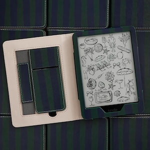 May include: A Kindle e-reader in a dark green and navy blue striped case. The interior of the case is light beige with a pocket. The screen displays a black and white illustration of various food items and beach objects with the text Sardines.