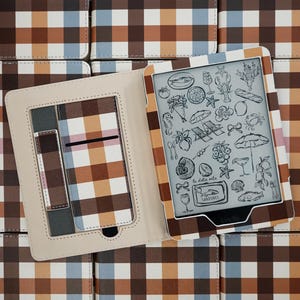 Op de afbeelding: Een e-reader in een beschermhoes met een bruin, beige, blauw en wit ruitpatroon. Het scherm toont een verzameling zwart-wit lijntekeningen van eten, drinken en strandartikelen met de tekst la dolce vita.