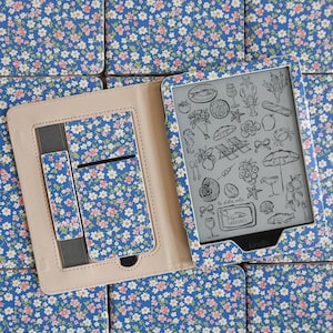 May include: A Kindle e-reader in a blue floral case with a beige interior. The screen displays a collection of black and white line drawings including a sardine tin labeled Sardines, la dolce vita, a lobster, beach chairs, and various fruits.