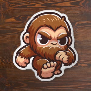 May include: A plush pillow shaped like a cartoon Bigfoot with brown fur, large dark eyes, and a determined expression. The character is depicted in a dynamic pose with one foot raised, featuring a white border outline against a dark wood background.