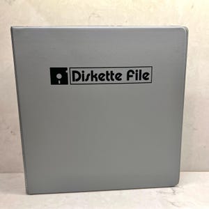 May include: A gray binder with the text "Diskette File" in a black font. The binder is rectangular and has a smooth surface. The text is inside a black rectangle.