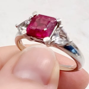 May include: A silver ring featuring a large, square-cut ruby gemstone as the centerpiece. Two smaller, triangular-shaped clear gemstones flank the ruby. The ring is held up, showcasing its design and the vibrant red color of the ruby.