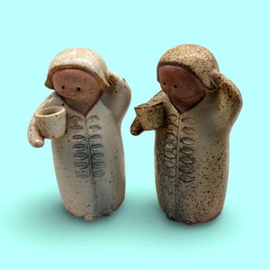 Może przedstawiać: Dwie ceramiczne figurki przedstawiające postacie w szatach z kapturami. Każda figurka ma nakrapiane wykończenie, gładką twarz z dwiema małymi kropkami zamiast oczu i trzyma w jednej dłoni mały kubek. Lewa figurka jest w kolorze jasnego kremu.