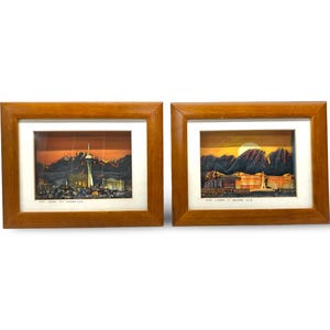 May include: Two framed landscape art pieces. One depicts a city skyline with a tall tower, the other a mountain range and buildings, both under a sunset sky. The frames are made of wood.