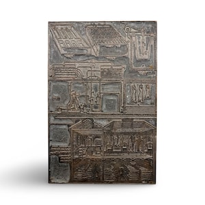 May include: A rectangular, gray metal printing plate with detailed relief carvings depicting architectural and industrial scenes. The plate features various structures, machinery, and figures, showcasing a complex design. The overall aesthetic is vintage and industrial.
