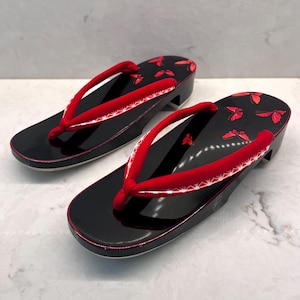 May include: A pair of black geta sandals with a glossy finish, featuring red fabric thong straps with white geometric patterns. The footbed is decorated with small red butterfly motifs, and the sandals are elevated on a solid black base.