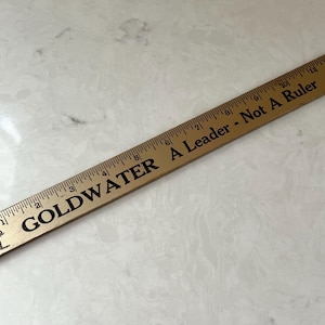 May include: A wooden ruler with black text that reads "GOLDWATER A Leader - Not A Ruler." The ruler has inch measurements and a small elephant graphic. The ruler is approximately 30 cm long.