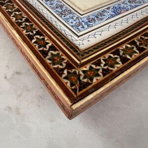 May include: A rectangular wooden box featuring intricate marquetry with geometric star patterns in green, red, and white. The lid displays a central miniature painting of a figure in a red garment, surrounded by a blue and white floral border.