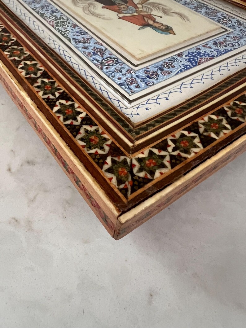 May include: A rectangular wooden box featuring intricate marquetry with geometric star patterns in green, red, and white. The lid displays a central miniature painting of a figure in a red garment, surrounded by a blue and white floral border.