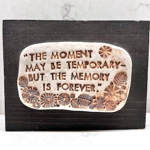 Op de afbeelding: Witte keramische plaquette met bruine bloemmotieven en de quote "The moment may be temporary - but the memory is forever."