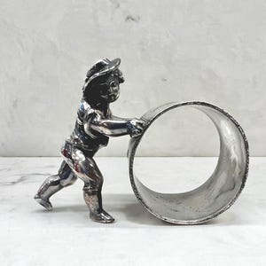 Op de afbeelding: Een zilverkleurige servetring met een gedetailleerd beeldje van een kind dat een grote ring duwt. Het kind draagt een hoed en kleding, met ingewikkelde details. De ring heeft een getextureerde rand, wat visuele interesse toevoegt aan dit decoratieve stuk.