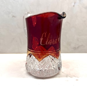 May include: A red glass pitcher with a clear glass base and gold trim. The pitcher has a handle and is decorated with a diamond pattern. The pitcher is inscribed with the name "Clara".