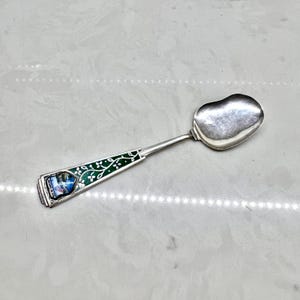 May include: A silver spoon with a heart-shaped bowl. The handle features a green enamel design with white floral accents and a small, colorful image. A decorative collectible.