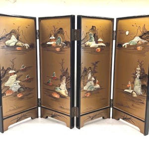 Puede incluir: Una pantalla plegable de cuatro paneles con fondo dorado y borde negro. Cada panel presenta una escena de paisaje con montañas, figuras y barcos. La pantalla está articulada y se mantiene en posición vertical, mostrando la obra de arte detallada.