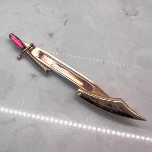 May include: A gold-toned metal brooch shaped like a sword or dagger. The handle features a vibrant, polished pink inlay, while the blade is long, slender, and pointed with a metallic finish. The design is sleek, elongated, and reflective.
