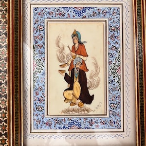 May include: A framed miniature painting featuring a seated figure in a dark robe and yellow trousers holding a bowl, with a standing figure in a red top and blue head covering behind, surrounded by a decorative border with blue floral patterns.
