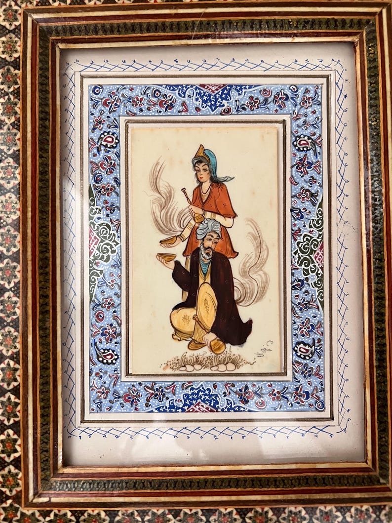May include: A framed miniature painting featuring a seated figure in a dark robe and yellow trousers holding a bowl, with a standing figure in a red top and blue head covering behind, surrounded by a decorative border with blue floral patterns.