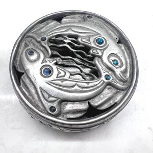 May include: A silver circular trinket box with a design of two stylized fish with blue gemstones for eyes. The box has a textured surface and a raised rim.
