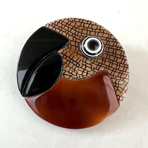 Puede incluir: Un objeto decorativo redondo con un acabado brillante. El diseño presenta una forma negra curva, una sección marrón y un área beige texturizada con un patrón de cuadrícula. También hay un pequeño detalle circular en blanco y negro.