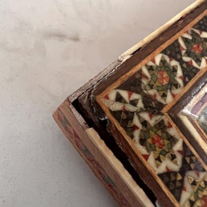 May include: A rectangular wooden box featuring intricate geometric inlay work on the lid. The surface displays a mosaic pattern of white, dark brown, green, and red triangular pieces arranged in star-like shapes, framed by dark wood borders.