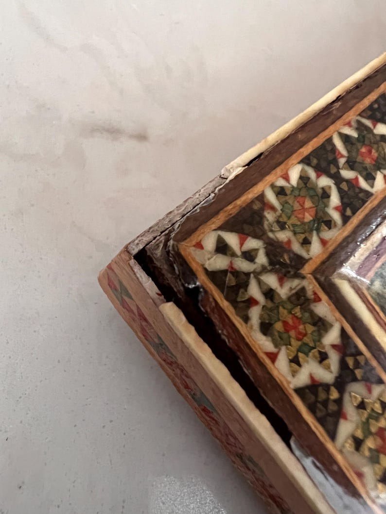 May include: A rectangular wooden box featuring intricate geometric inlay work on the lid. The surface displays a mosaic pattern of white, dark brown, green, and red triangular pieces arranged in star-like shapes, framed by dark wood borders.