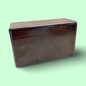 May include: A rectangular, dark brown box with a slightly glossy finish. The box has a lid and appears to be made of a solid material. The edges are rounded, and the overall design is simple and classic. The box is set against a light green background.