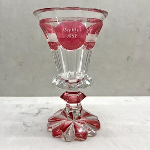 Puede incluir: Una copa de cristal transparente con detalles en rojo. La copa tiene un borde acampanado y una base facetada. El texto "Teplitz. 1844." es visible en la copa. La copa es probablemente vintage o antigua.