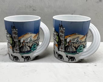 May include: Two white ceramic mugs with a blue gradient background featuring a detailed illustration of a cityscape with a bridge, buildings, and a dome. The mugs have white, curved handles and a repeating pattern of black animal figures.