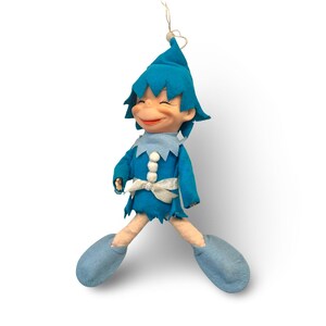 Puede incluir: Una muñeca vintage de fieltro azul con una cara sonriente, que lleva un sombrero, una túnica y zapatos a juego. La muñeca tiene un lazo blanco y un cuello azul claro. La muñeca está suspendida por un hilo dorado, lo que sugiere que es un adorno.