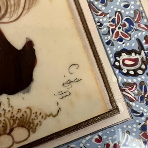 May include: A rectangular painting on a light-colored surface featuring a dark brown figure and intricate gold-colored brushwork at the bottom. The artwork is framed by a border with a light blue background, white dots, and red and blue floral motifs.