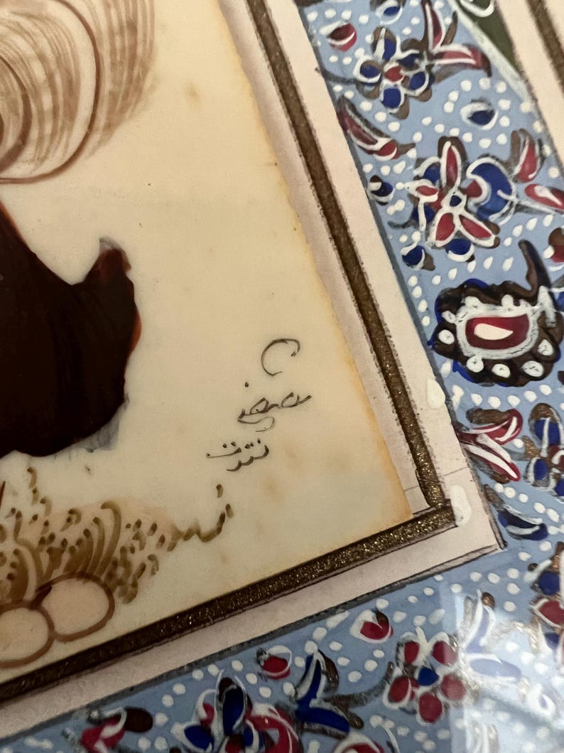 May include: A rectangular painting on a light-colored surface featuring a dark brown figure and intricate gold-colored brushwork at the bottom. The artwork is framed by a border with a light blue background, white dots, and red and blue floral motifs.