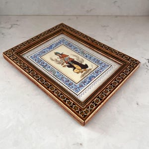 May include: A rectangular framed painting featuring a central illustration of a figure in traditional attire, surrounded by a blue and white floral border. The frame is made of wood with a dark brown finish and intricate geometric star patterns.