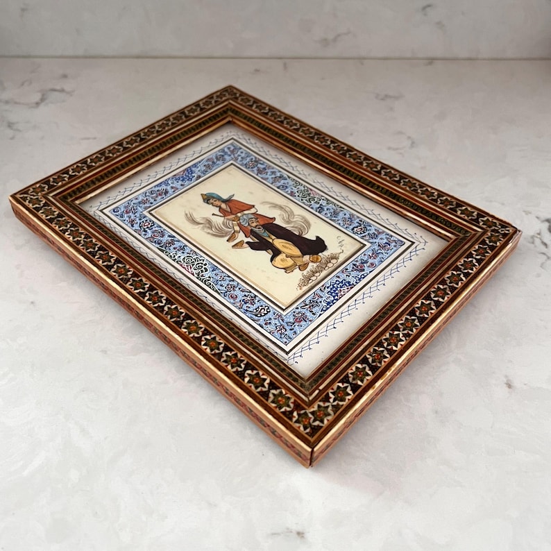 May include: A rectangular framed painting featuring a central illustration of a figure in traditional attire, surrounded by a blue and white floral border. The frame is made of wood with a dark brown finish and intricate geometric star patterns.