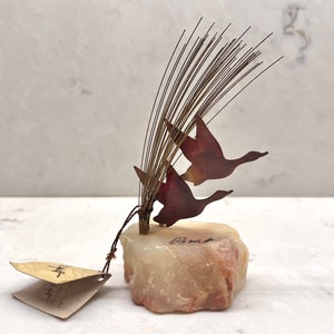 May include: A metal sculpture of two geese in flight, with a long, thin, metal tail. The sculpture is mounted on a small, pink, natural stone base. The sculpture is signed "Ed. No. 1".