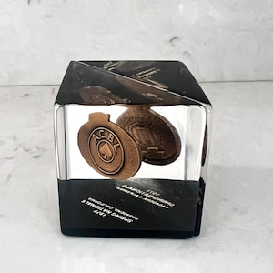 Può includere: Un fermacarte a forma di cubo trasparente con un emblema color bronzo con le lettere "A.C.B.L." e un simbolo di picche. La base nera presenta il testo bianco: "1977 Spring Nationals Pasadena, California".