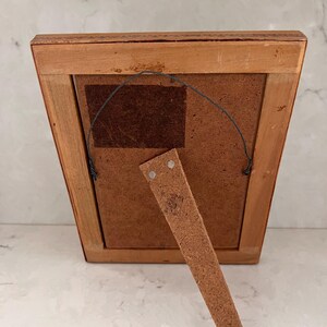 May include: The back of a rectangular wooden picture frame featuring a light brown finish, a dark brown fiberboard backing, a metal hanging wire attached to two small eyelets, and a hinged wooden support stand extending from the lower center.