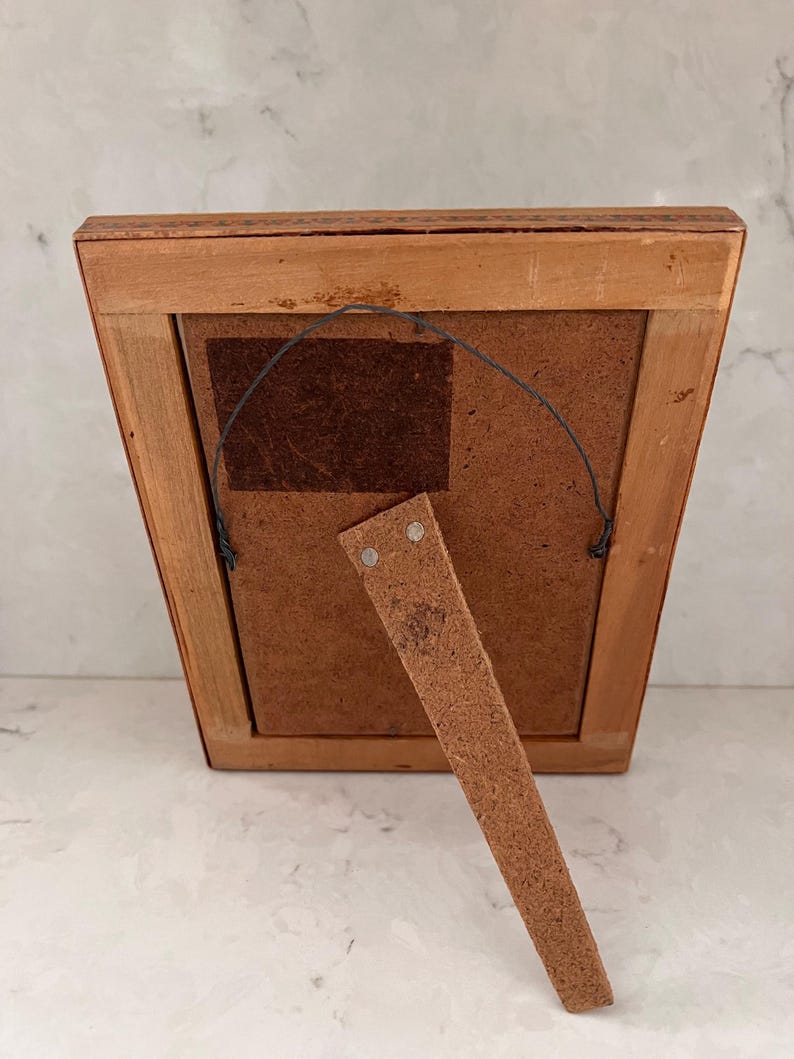 May include: The back of a rectangular wooden picture frame featuring a light brown finish, a dark brown fiberboard backing, a metal hanging wire attached to two small eyelets, and a hinged wooden support stand extending from the lower center.