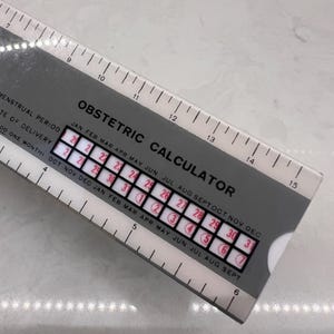 May include: A gray obstetric calculator with a white ruler edge measuring up to 15 inches. The device features black text including "OBSTETRIC CALCULATOR," "MENSTRUAL PERIOD," and "DATE OF DELIVERY," with a sliding window showing red numbers.