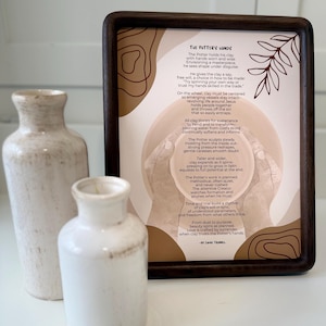 May include: A framed print with the text "The Potter's Hands" and a poem, alongside two white ceramic vases. The print has a brown frame and a beige background with abstract shapes. The vases are different sizes and textures.