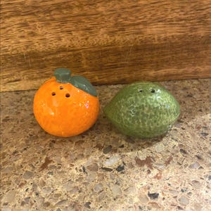 May include: A pair of ceramic salt and pepper shakers shaped like a bright orange and a green lime. The orange shaker has a green stem and leaf, and the lime shaker has a textured surface.