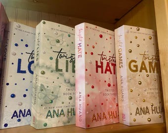 May include: Four books from the "Twisted" series by Ana Huang are displayed on a shelf. The spines are white, pink, and gold, decorated with beads. Titles include "Twisted Love," "Twisted Lies," "Twisted Hate," and "Twisted Games."
