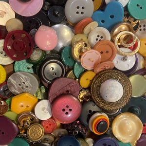 May include: A collection of various buttons in different sizes, shapes, and colors. The buttons include shades of pink, black, gray, yellow, and blue. Some buttons have four holes, while others have two. There are also gold and iridescent buttons.